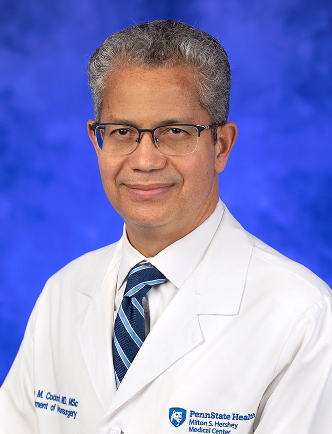 Kevin M. Cockroft, MD, MSc, FAANS, FACS, FAHA, is Interim Chair of the Department of Neurosurgery at Penn State College of Medicine. He is pictured in a white medical coat against a blue background.